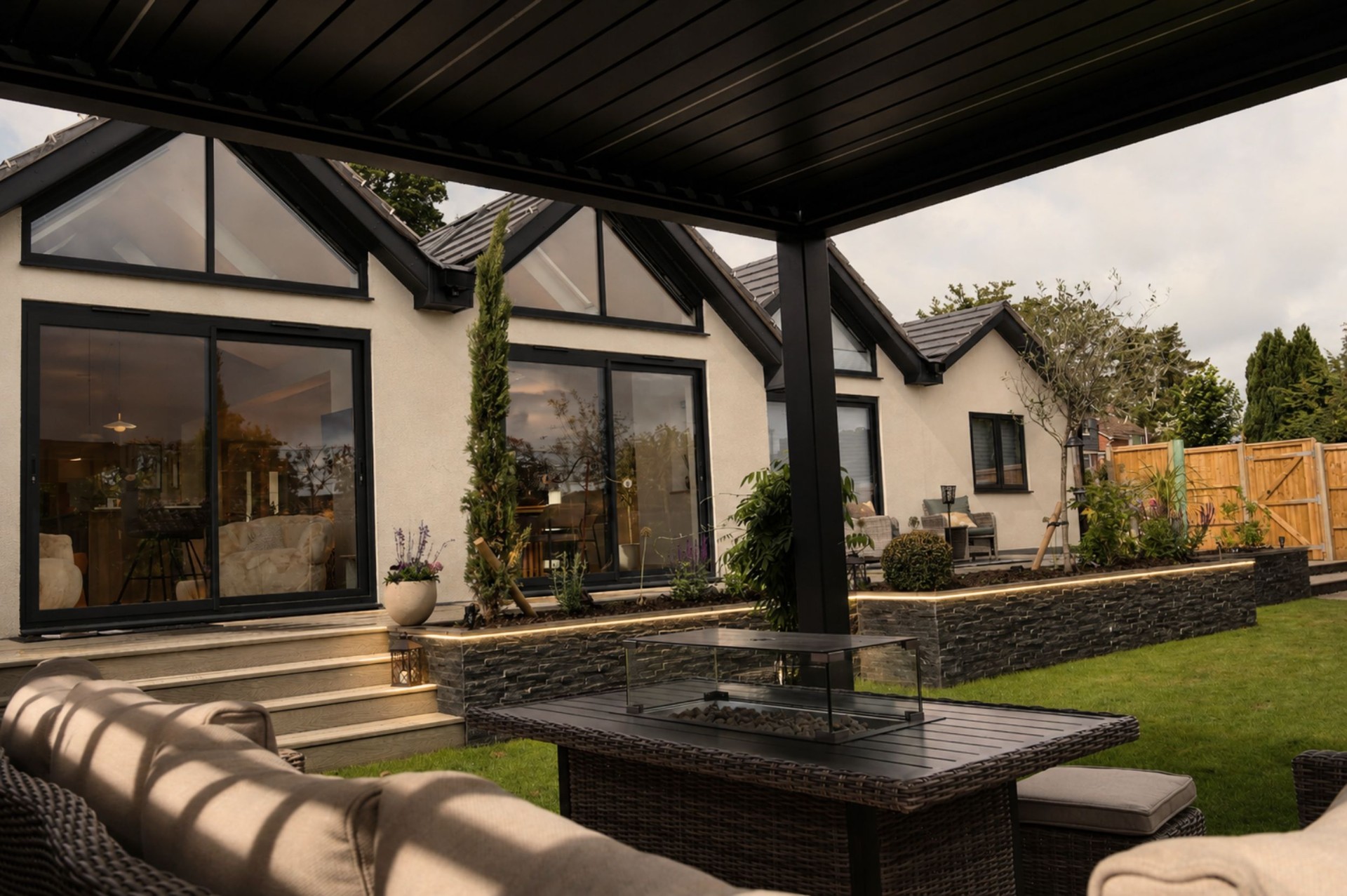 Charcoal louvered pergola over a deck with fire-pit lounge, stone-clad raised beds and integrated LED strip lighting