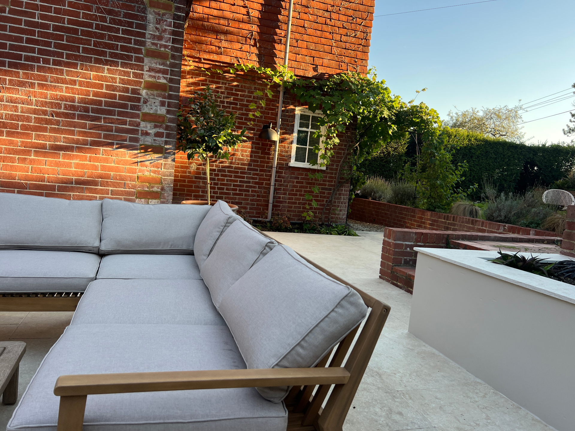 Medi Garden — travertine terrace with a low grey sofa beneath a vine-clad brick wall at golden hour