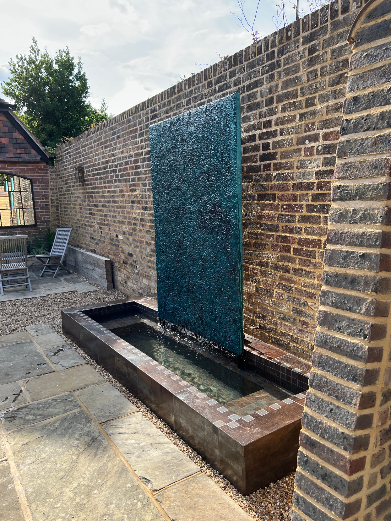 Tall lava-stone water wall cascading into a copper-tiled sunken rill beneath the brick boundary wall