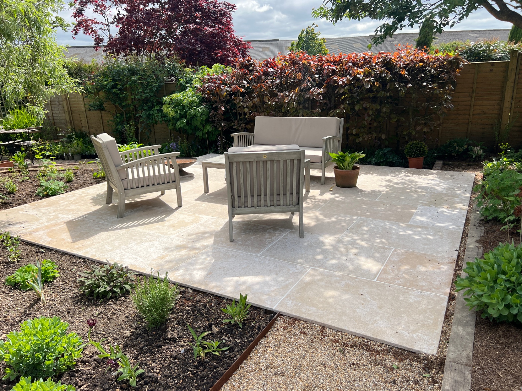 Square limestone patio with teak lounge set, fire bowl and copper beech hedge backdrop